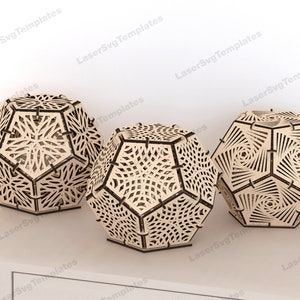 May include: Three wooden geometric lampshades with intricate laser cut designs. The lampshades are in the shape of a dodecahedron and are made of light brown wood.