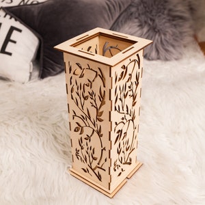 Pode incluir: Um vaso decorativo de madeira com um design floral. O vaso &eacute; de cor castanho claro e tem uma forma quadrada. Est&aacute; sentado sobre uma superf&iacute;cie branca e fofa.