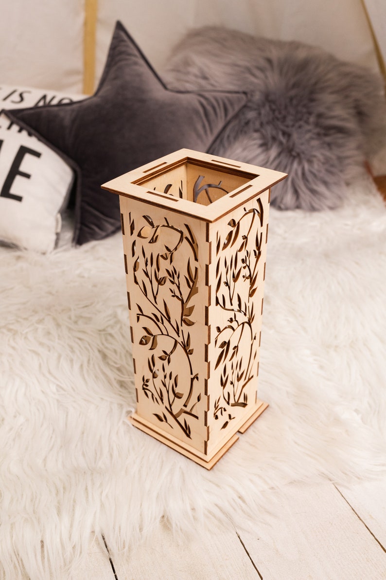 Pode incluir: Um vaso decorativo de madeira com um design floral. O vaso &eacute; de cor castanho claro e tem uma forma quadrada. Est&aacute; sentado sobre uma superf&iacute;cie branca e fofa.