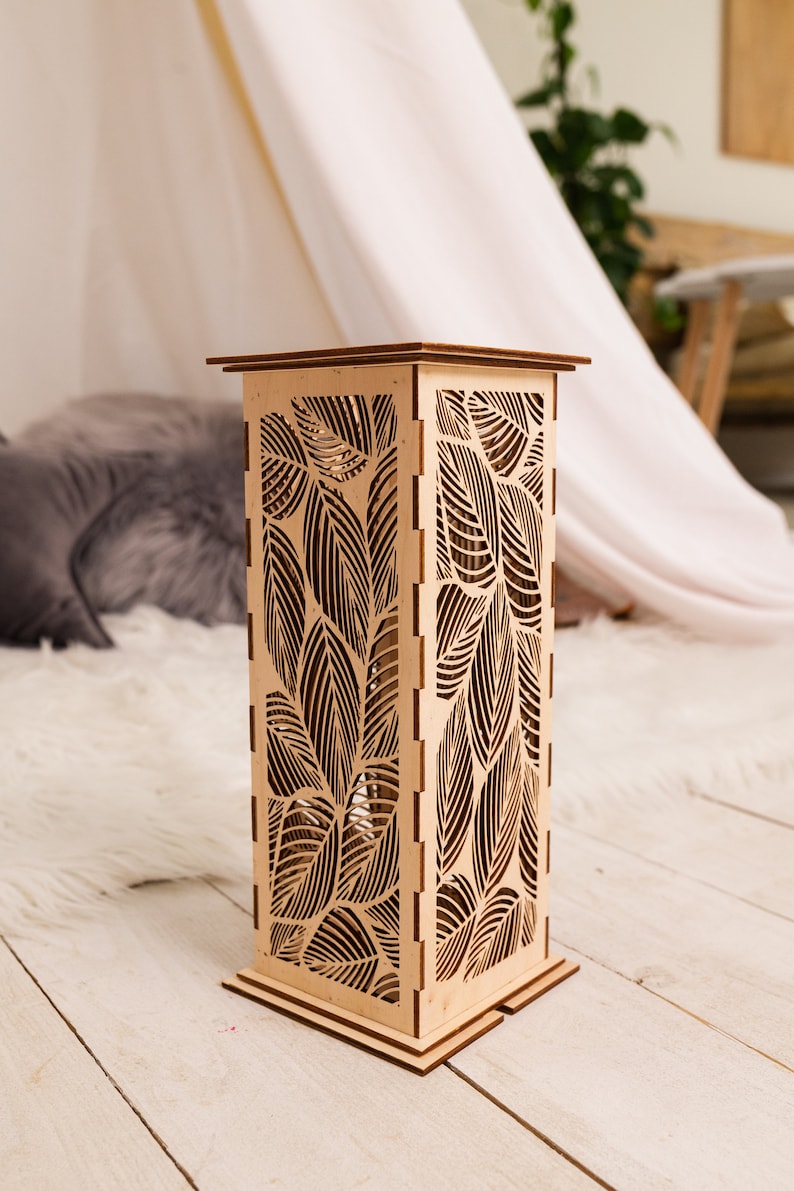 Pode incluir: Uma l&acirc;mpada de madeira com um design de folhas. A l&acirc;mpada &eacute; feita de madeira marrom claro e tem uma base quadrada. As folhas s&atilde;o cortadas na madeira e criam um padr&atilde;o delicado.