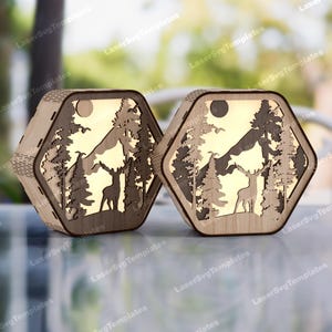Könnte beinhalten: Zwei sechseckige Holzlaternen mit ausgeschnittener Waldszene. Das Design zeigt einen Hirsch, Bäume, Berge und einen Mond. Die Laternen sind aus hellem Holz gefertigt und haben einen warmen Schein von innen.