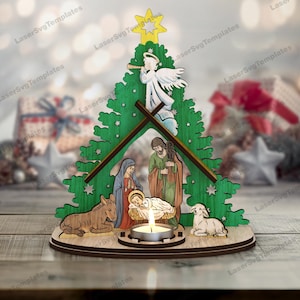May include: A wooden Christmas nativity scene with a green tree-shaped backdrop. The scene includes figures of Mary, Joseph, baby Jesus, an angel, a donkey, and a lamb. A lit candle sits in front of the figures. The text "LaserSvg Templates" is visible.