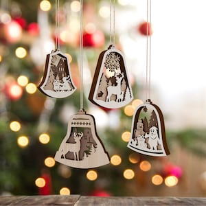Puede incluir: Cuatro adornos navideños de madera con forma de campana con intrincados diseños recortados. Los adornos presentan escenas de invierno con un ciervo, un muñeco de nieve, una casa y un copo de nieve.