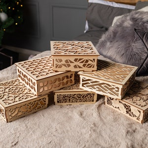 May include: Six wooden boxes with intricate laser-cut designs stacked on a beige surface. The boxes are made of light brown wood and feature various geometric and floral patterns.