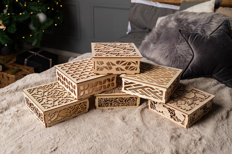 May include: Six wooden boxes with intricate laser-cut designs stacked on a beige surface. The boxes are made of light brown wood and feature various geometric and floral patterns.