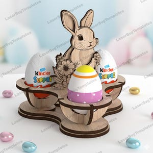 Może przedstawiać: Drewniany stojak na pisanki z sylwetką królika i trzema wycięciami w kształcie jajek. Wyświetlane są dwa jajka Kinder Niespodzianka i dekoracyjne jajko w różowe grochy. Stojak otoczony jest pastelowymi pisankami.