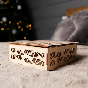 May include: A wooden box with a geometric design, sitting on a white fuzzy surface. The box is made of light wood and has a delicate, intricate pattern.