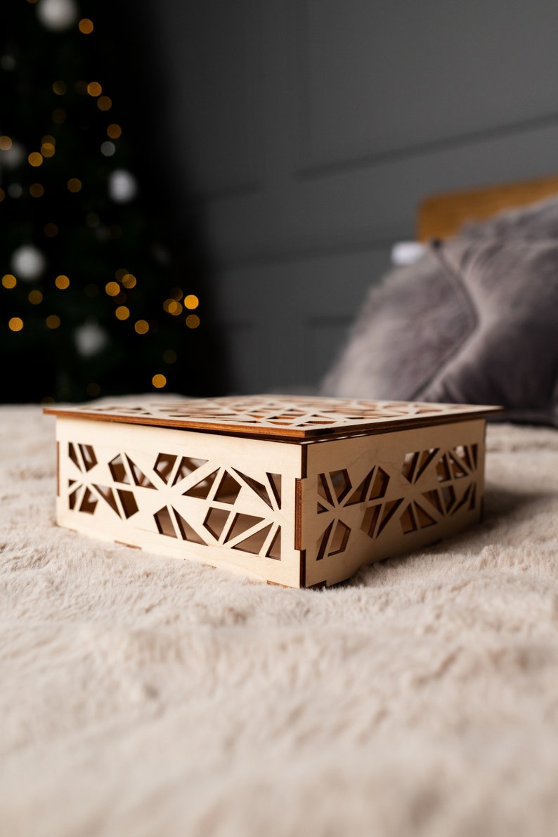 May include: A wooden box with a geometric design, sitting on a white fuzzy surface. The box is made of light wood and has a delicate, intricate pattern.