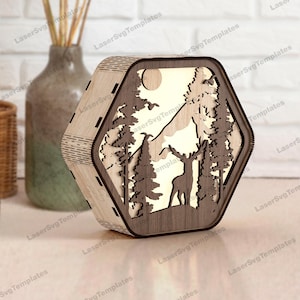 Puede incluir: Caja de luz hexagonal de madera con una escena de bosque cortada con láser que presenta un ciervo, árboles, montañas y una luna. La caja tiene un acabado de madera natural y un brillo cálido.