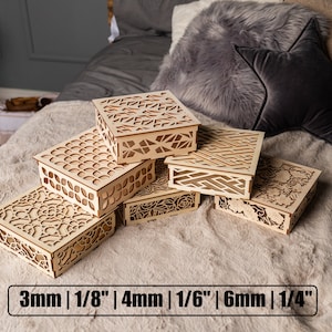 May include: Five wooden boxes with intricate laser-cut designs, stacked on a light brown faux fur surface. The boxes are made of natural wood and have a variety of patterns, including geometric shapes, flowers, and swirls. The boxes are labeled with their thickness in millimeters and inches: 3mm | 1/8", 4mm | 1/6", 6mm | 1/4".