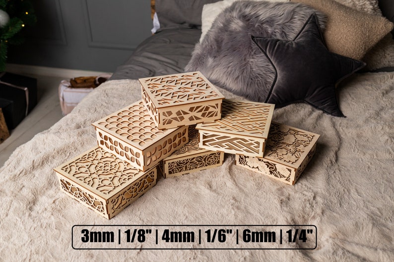 May include: Five wooden boxes with intricate laser-cut designs, stacked on a light brown faux fur surface. The boxes are made of natural wood and have a variety of patterns, including geometric shapes, flowers, and swirls. The boxes are labeled with their thickness in millimeters and inches: 3mm | 1/8", 4mm | 1/6", 6mm | 1/4".