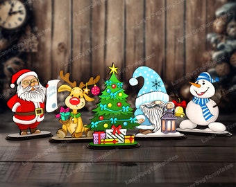 Op de afbeelding: Een set van vijf kerstthema houten uitsnijdingen. De uitsnijdingen omvatten de kerstman die een rol houdt met de tekst "Merry Christmas", een rendier dat een cadeau vasthoudt, een kerstboom met cadeaus, een kabouter die een lantaarn vasthoudt en een sneeuwpop met een blauwe sjaal.