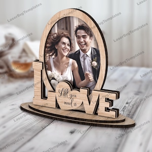 May include: Wooden picture frame with the word "LOVE" in large letters and a heart-shaped cutout that says "All you need is". The frame is designed to hold a photo of a couple.