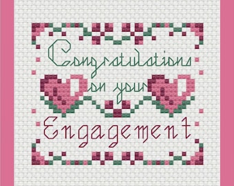 Puede incluir: Un patrón de punto de cruz sobre tela Aida blanca con un borde rosa. El texto dice Congratulations on your Engagement en hilo verde oscuro y granate. Dos corazones rosas pixelados están centrados entre las palabras, rodeados de motivos florales.