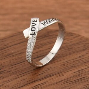 May include: A silver-toned ring featuring a crossover design with the word LOVE engraved on one end and a row of small clear stones embedded along the band. The metal surface is polished and reflective, showcasing a clean and simple aesthetic.