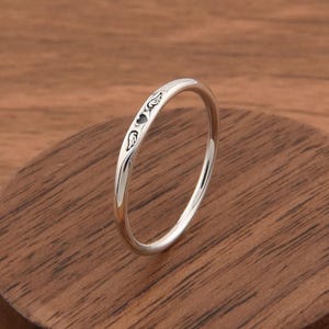 May include: A thin, polished silver ring featuring a small, engraved heart symbol flanked by two delicate wing motifs on its outer surface. The band has a smooth, rounded profile and a reflective metallic finish, showcasing fine, detailed line work.