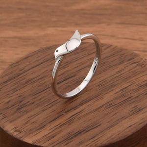 May include: A silver ring featuring a bird design. The ring is displayed on a wooden block with a dark brown finish. The bird's head and body are clearly defined, with a smooth, polished surface. The ring's band is thin and circular.