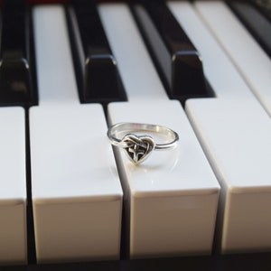 May include: A silver ring with a heart-shaped design featuring a cross inside. The ring is resting on a white surface.