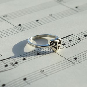 May include: A silver ring with a heart-shaped design featuring a Celtic knot and a cross. The ring is lying on a sheet of music.