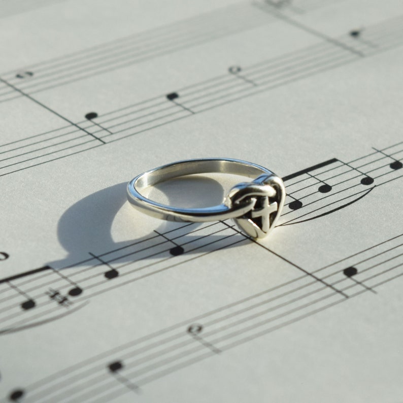 May include: A silver ring with a heart-shaped design featuring a Celtic knot and a cross. The ring is lying on a sheet of music.