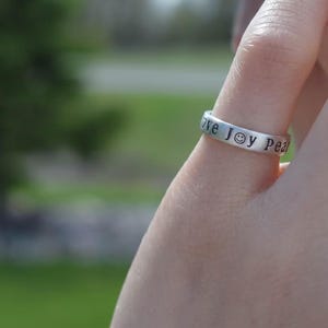 May include: A silver ring engraved with the words "love joy peace" around the band. The word "joy" is represented by a smiley face. The ring is on a finger, with a blurred green background.