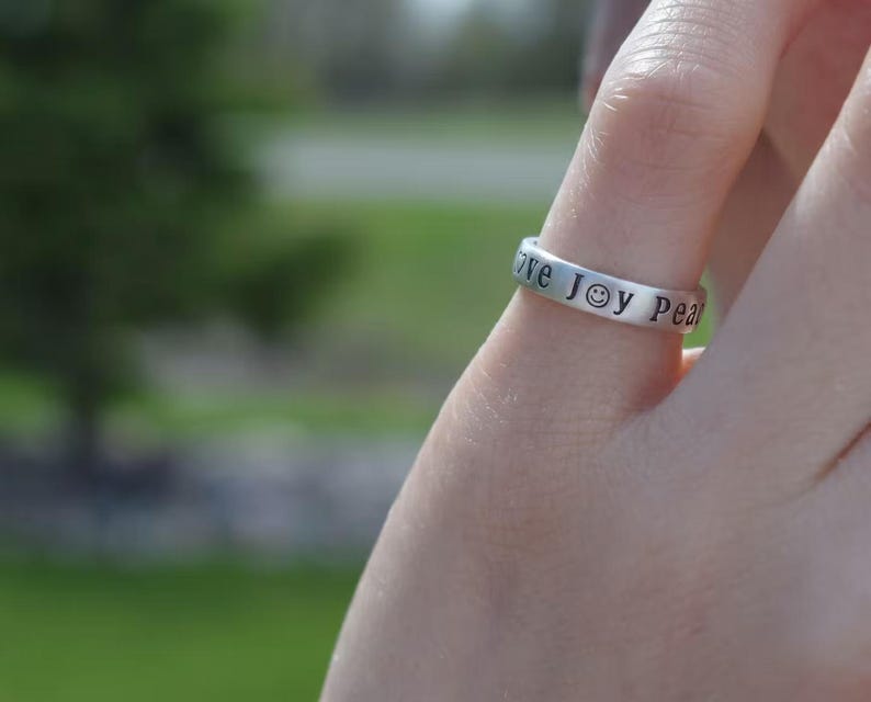 May include: A silver ring engraved with the words "love joy peace" around the band. The word "joy" is represented by a smiley face. The ring is on a finger, with a blurred green background.