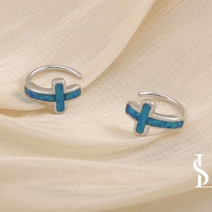 May include: Two silver-toned cross-shaped earrings with blue opal inlays. The earrings are set against a cream-colored fabric background. The cross design is a religious symbol.