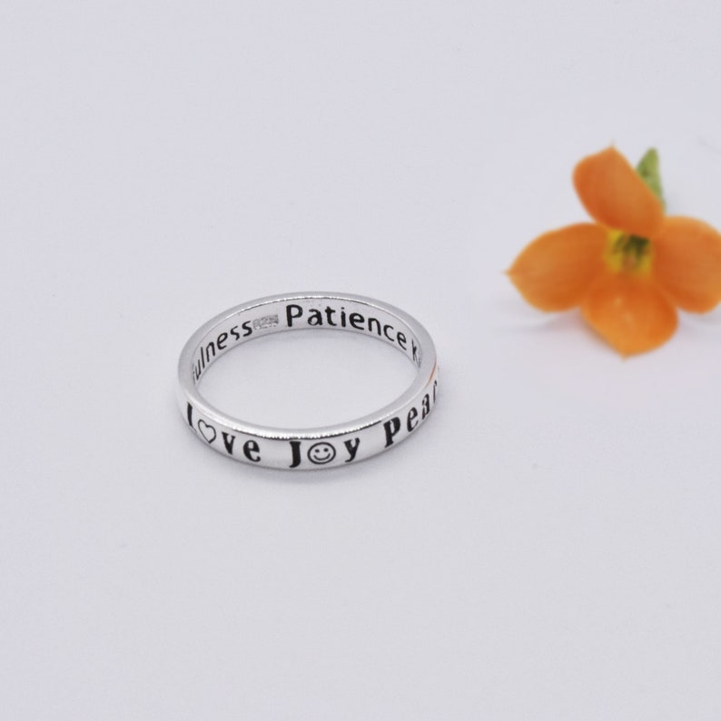 May include: A silver ring engraved with the words "love", "joy", "peace", and "patience". The ring also features a heart and a smiley face symbol. An orange flower is in the background.