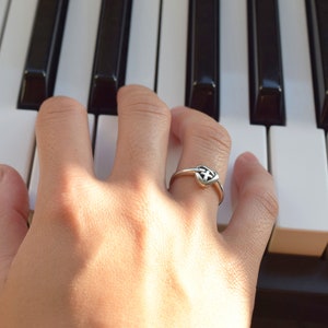 May include: A silver ring with a heart-shaped design featuring a cross inside. The ring is worn on the finger of a hand resting on a piano keyboard.