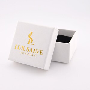 May include: A white jewellery box with the text "LUX SALVE JEWELRY" in gold lettering. The box is open, revealing a black interior. The box is on a white surface.