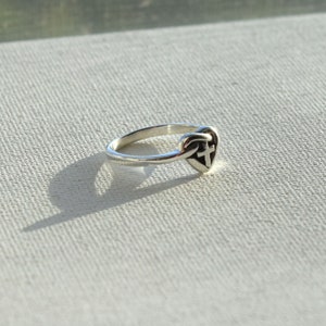 May include: A silver ring with a heart-shaped design featuring a cross inside. The ring is on a white textured surface.