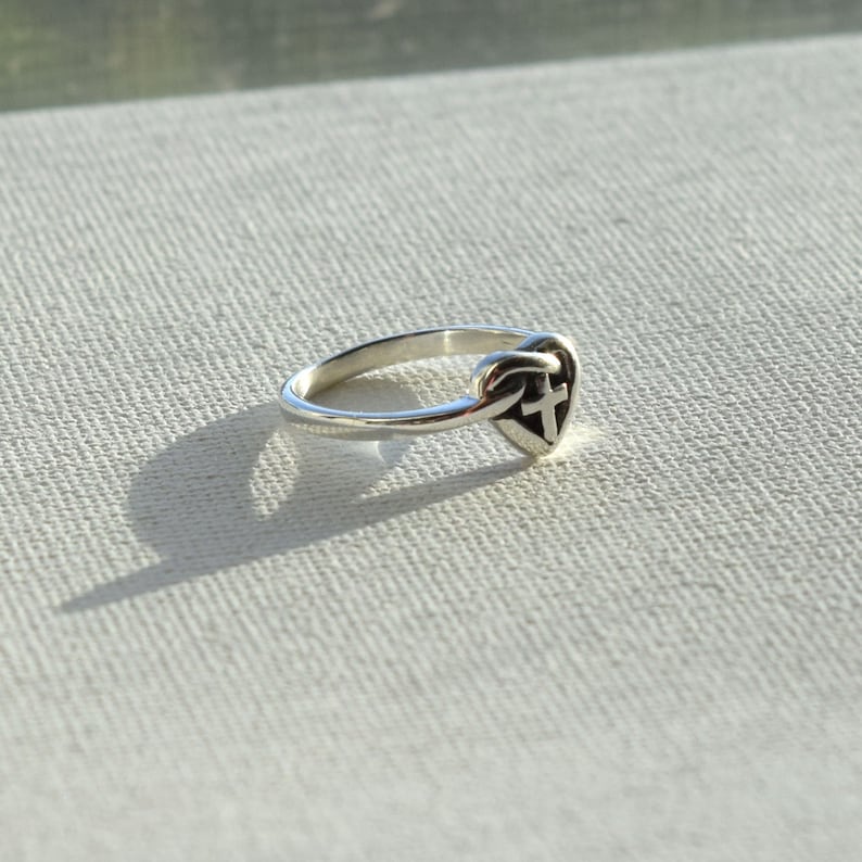 May include: A silver ring with a heart-shaped design featuring a cross inside. The ring is on a white textured surface.