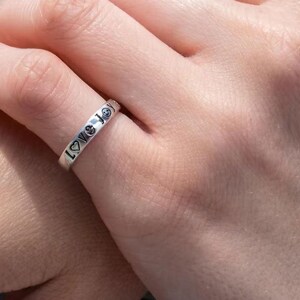 May include: A silver ring engraved with the words "Love Joy" and smiley face and heart symbols. The ring is worn on a finger.