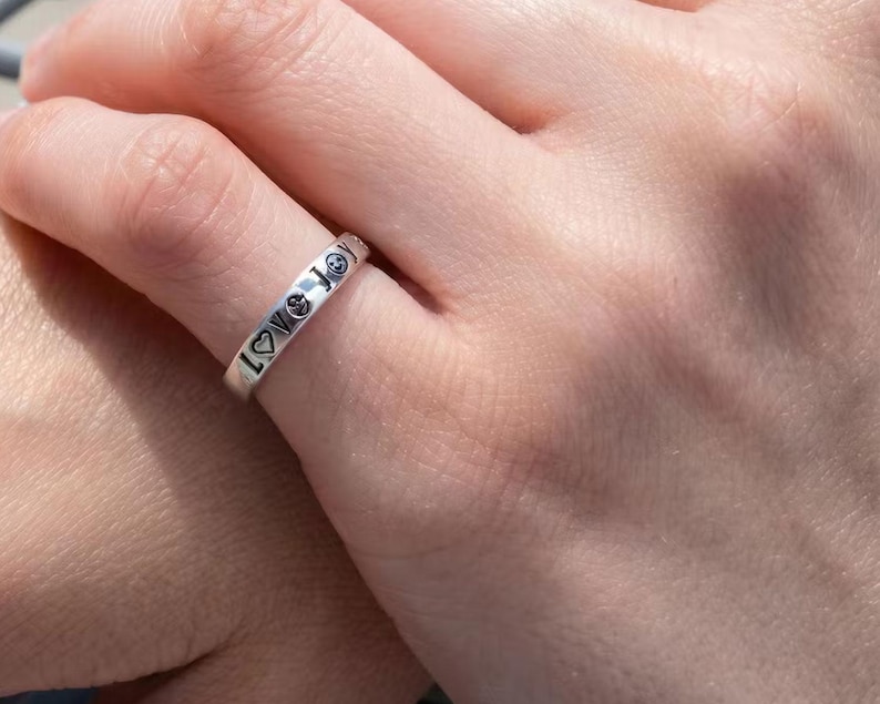 May include: A silver ring engraved with the words "Love Joy" and smiley face and heart symbols. The ring is worn on a finger.