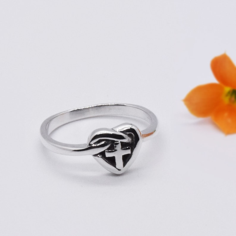 May include: A silver ring with a heart-shaped design featuring a cross inside. The ring is on a white background.