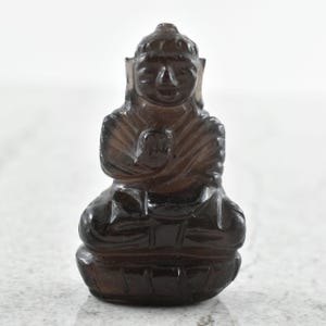 May include: A small, translucent, dark brown smoky quartz carving of a seated figure in a meditative pose. The figure features carved facial details, draped robes with visible line textures, and sits upon a stylized, rounded base.