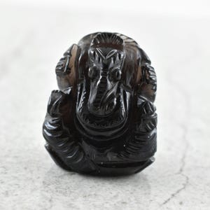 May include: A dark, translucent smoky quartz carving of the deity Ganesha. The figure is depicted in a seated position with a prominent trunk, large ears, and detailed facial features, showcasing the natural deep brown and black tones of the stone.
