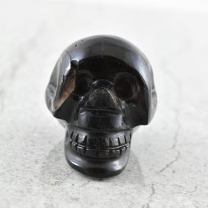 May include: A polished black stone carving shaped like a human skull. The object features deep-set eye sockets, a defined nasal cavity, and carved teeth along the jawline. A visible natural fissure runs vertically along the left side of the cranium.