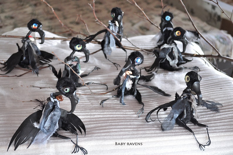 K&ouml;nnte beinhalten: Eine Gruppe von acht schwarzen Rabenfiguren mit wei&szlig;en und blauen Stoffakzenten. Die Raben haben gro&szlig;e, bunte Augen und sind auf einer wei&szlig;en Oberfl&auml;che mit &Auml;sten angeordnet.