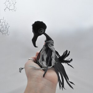 K&ouml;nnte beinhalten: Eine schwarze und graue Stoff-Rabenpuppe mit einem zerfetzten Stoffscarf. Die Puppe wird in der Hand einer Person gehalten.