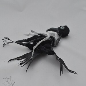 K&ouml;nnte beinhalten: Eine schwarze Rabenfigur mit einem wei&szlig;en Stofffl&uuml;gel, die auf einer wei&szlig;en Oberfl&auml;che liegt. Der Rabe hat ein einzelnes gr&uuml;nes Auge und einen schwarzen Schnabel.