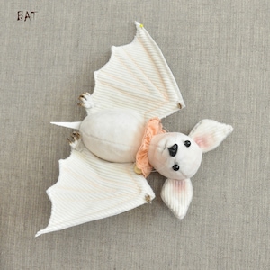 Könnte beinhalten: Eine weiße Plüschfledermaus mit rosa Ohren und einem rosa Rüschenkragen. Die Fledermaus hat schwarze Augen und eine kleine schwarze Nase. Die Fledermaus liegt auf dem Rücken mit ausgebreiteten Flügeln.
