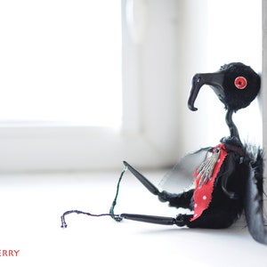 K&ouml;nnte beinhalten: Eine schwarze vogelartige Puppe mit roten Augen und einem rot-wei&szlig; gepunkteten Schal. Die Puppe hat schwarze Fl&uuml;gel und sitzt auf einer wei&szlig;en Oberfl&auml;che.