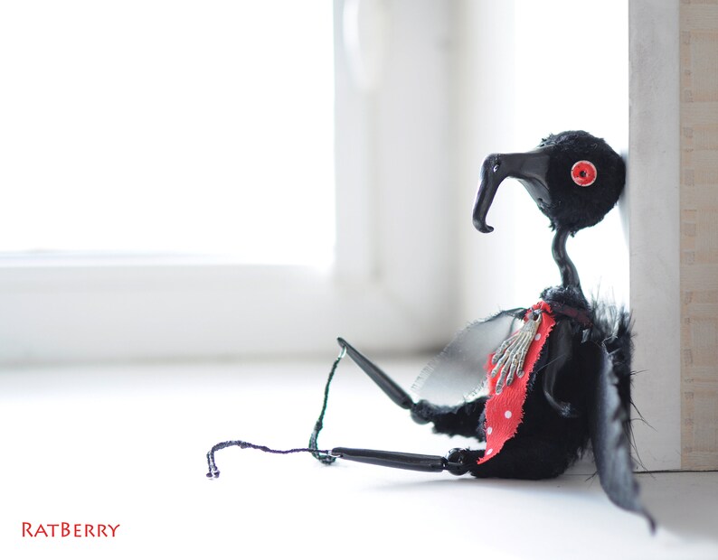 K&ouml;nnte beinhalten: Eine schwarze vogelartige Puppe mit roten Augen und einem rot-wei&szlig; gepunkteten Schal. Die Puppe hat schwarze Fl&uuml;gel und sitzt auf einer wei&szlig;en Oberfl&auml;che.