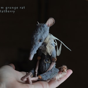 May include: A small, gray, handmade rat plush toy with a white lace collar and a heart-shaped pendant. The rat is sitting on a hand.