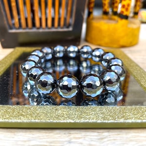 May include: A dark grey hematite bead bracelet with faceted surfaces. The beads are strung together, reflecting light. The bracelet is displayed on a mirrored surface with a gold-coloured frame.
