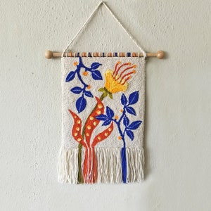 May include: A white wall hanging with a floral design. The design features a yellow flower with orange and blue leaves. The wall hanging has a wooden dowel at the top and a fringe at the bottom.