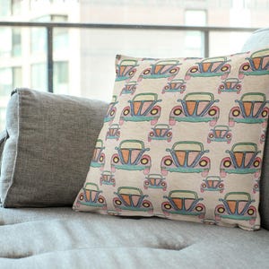 May include: A square cushion featuring a repeating pattern of vintage cars in blue, orange, and pink on a beige background. The cushion is placed on a grey sofa. The cars are reminiscent of classic vehicles.