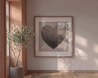 May include: A framed artwork featuring a textured, dark heart shape. The artwork is displayed in a room with a potted olive tree and a window. The room has a neutral color palette with warm tones and natural light.