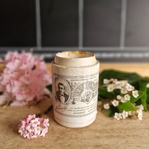 May include: A vintage ceramic jar with a chipped white glaze and a black and white label that reads "La Reine des Blemy" with a portrait of a man in a suit and bow tie. The label also includes the text "Diplome d'Honneur Paris" and "La seule sans rouille", and "Entrepôt S. Just."